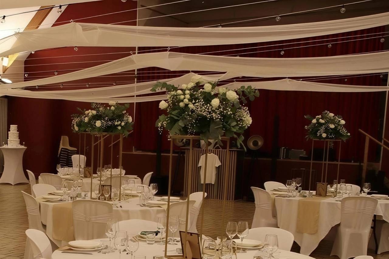 Salle de réception décorée de voilages ivoire, guirlandes lumineuses et grandes compositions de fleurs blanches sur supports dorés